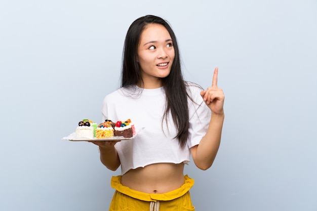 Teenager asian girl holding lots of different mini cakes intending to realizes the solution while lifting a finger up