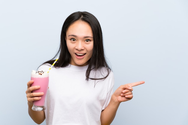 Teenager asian girl holding a strawberry milkshake surprised and pointing finger to the side