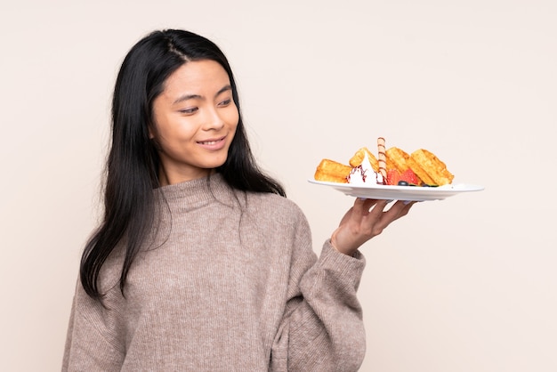 Teenager Asian girl holding waffles on beige wall with happy expression