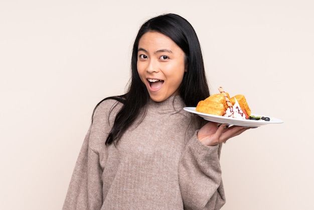 Teenager Asian girl holding waffles on beige wall with surprise and shocked facial expression