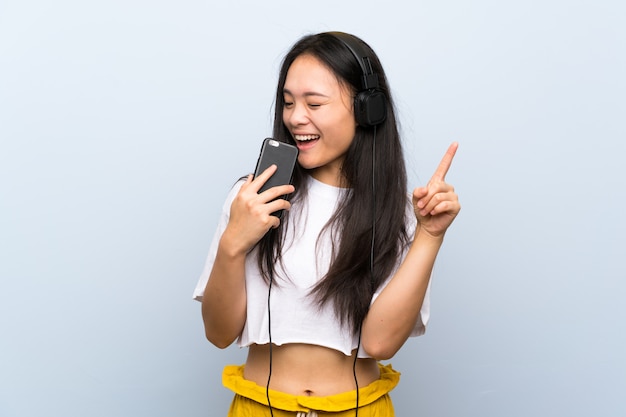 Teenager asian girl listening music over isolated blue wall singing