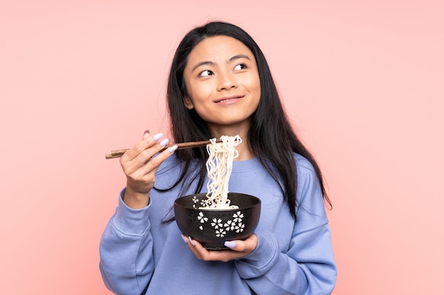 Teenager Asian woman on beige wall holding a bowl of noodles with chopsticks
