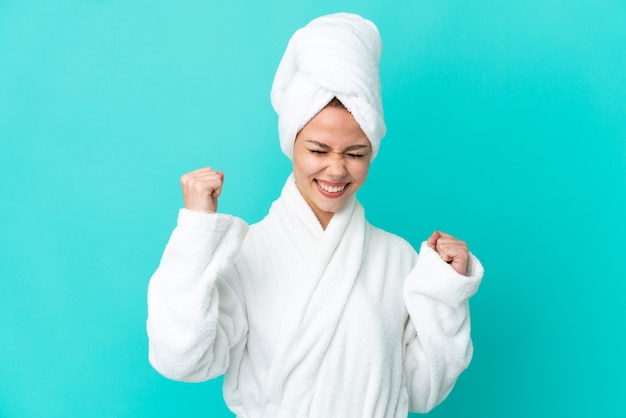 Teenager blonde girl in a bathrobe over isolated blue background celebrating a victory