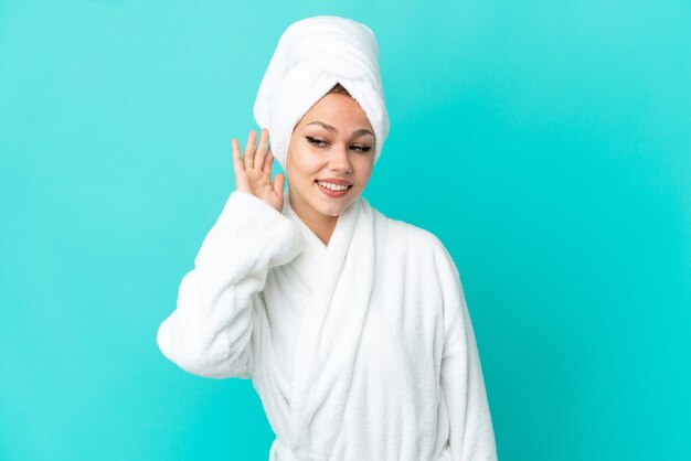 Teenager blonde girl in a bathrobe over isolated blue background listening to something by putting hand on the ear