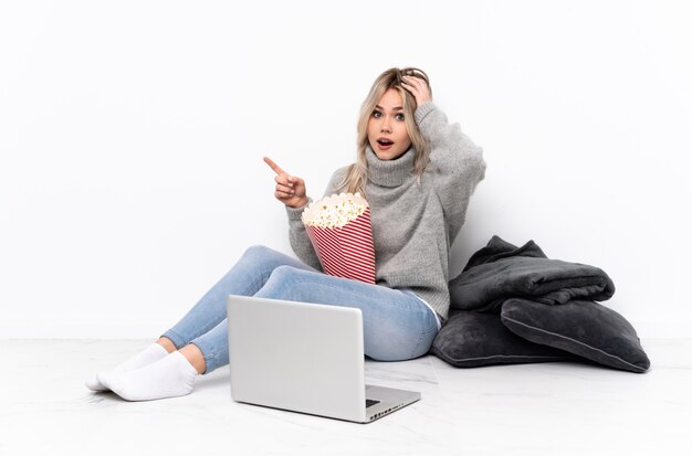 Teenager blonde girl eating popcorn while watching a movie on the laptop surprised and pointing finger to the side