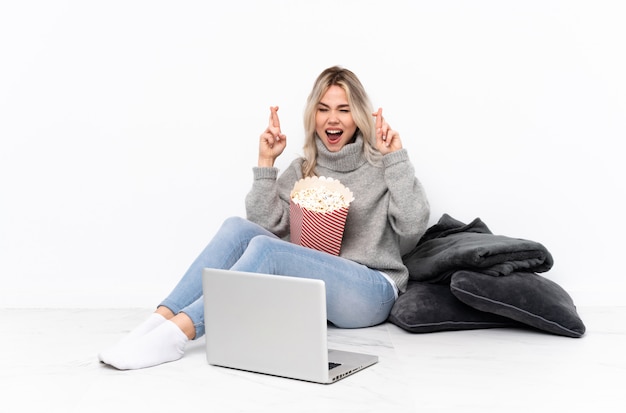Блондинка подросток ест попкорн во время просмотра фильма на ноутбуке с скрестив пальцы