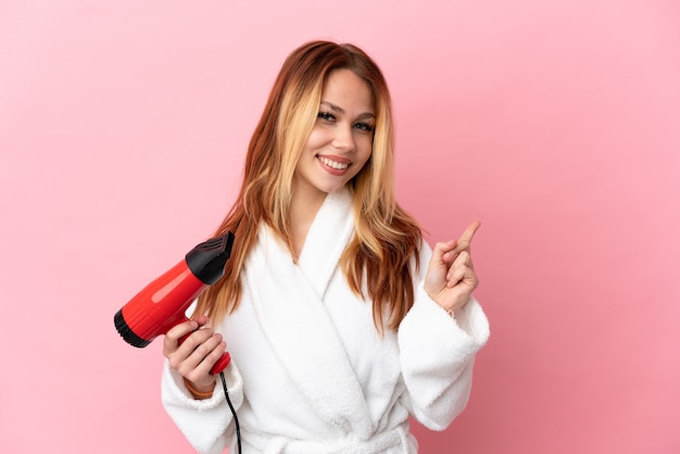 Teenager blonde girl holding a hairdryer over isolated pink background pointing back