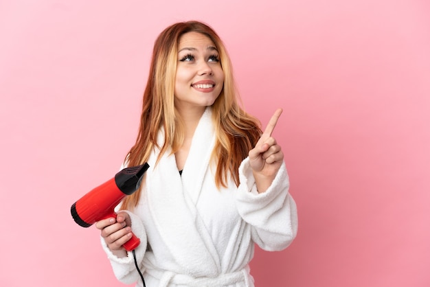 素晴らしいアイデアを指している孤立したピンクの背景の上にヘアドライヤーを保持しているティーンエイジャーのブロンドの女の子