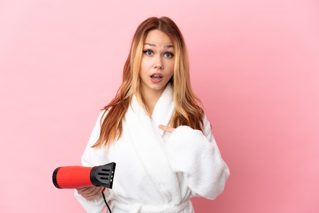 Teenager blonde girl holding a hairdryer over isolated pink background with surprise facial expression