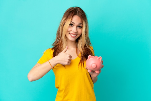 Teenager blonde girl holding a piggybank over isolated blue background giving a thumbs up gesture