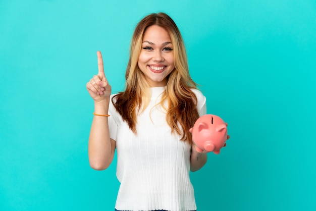 Teenager blonde girl holding a piggybank over isolated blue background pointing up a great idea