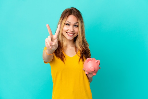 Teenager blonde girl holding a piggybank over isolated blue background smiling and showing victory sign