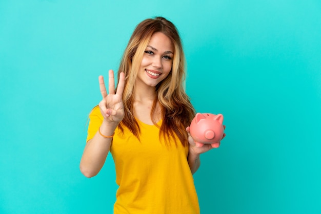 Teenager blonde girl holding a piggybank over isolated blue surface happy and counting three with fingers