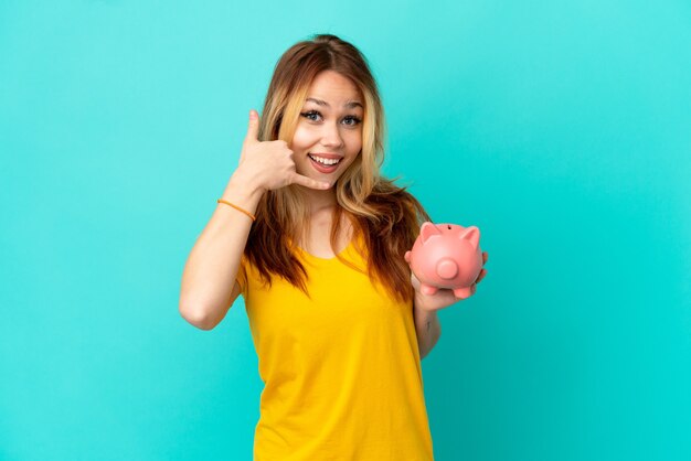 Teenager blonde girl holding a piggybank over isolated blue wall making phone gesture