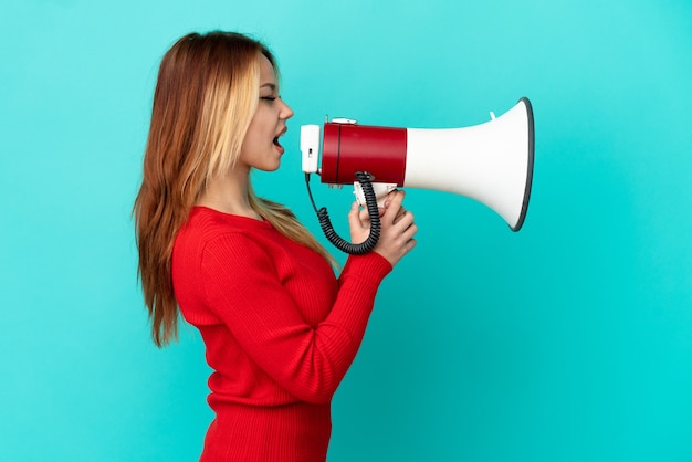 Teenager blonde girl over isolated blue background shouting through a megaphone to announce something in lateral position