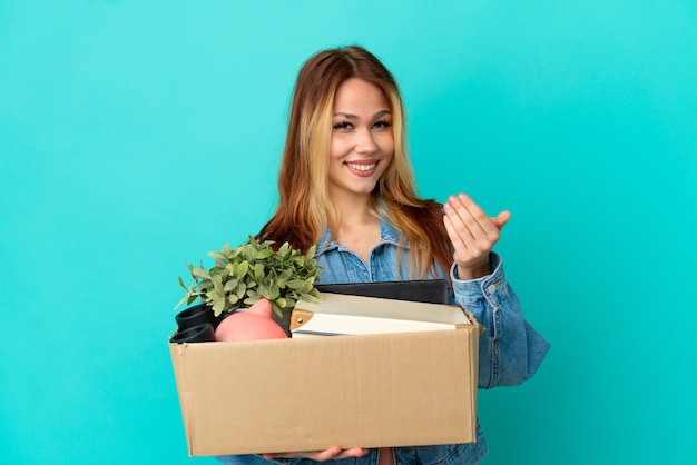 Teenager blonde girl making a move while picking up a box full of things inviting to come with hand. Happy that you came