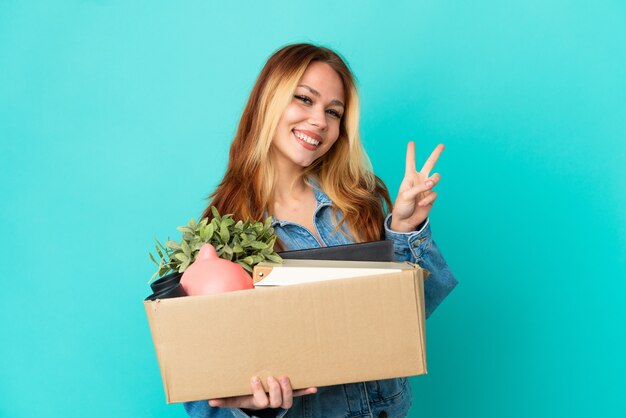 Teenager blonde girl making a move while picking up a box full of things smiling and showing victory sign