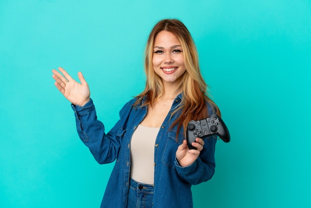 Adolescente ragazza bionda che gioca con un controller per videogiochi su un muro isolato che estende le mani di lato per invitare a venire