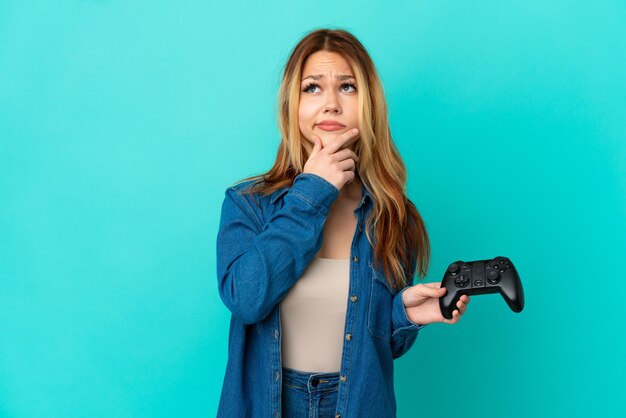 Teenager blonde girl playing with a video game controller over isolated wall having doubts