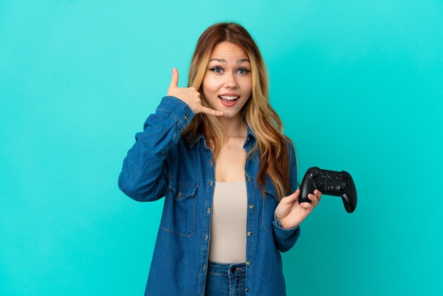 Девушка-подросток блондинка играет с контроллером видеоигры над изолированной стеной, делая телефонный жест. Перезвони мне знак
