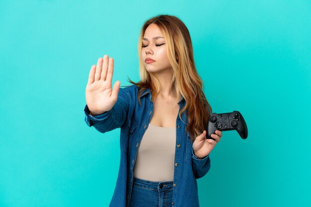 Teenager blonde girl playing with a video game controller over\
isolated wall making stop gesture and disappointed