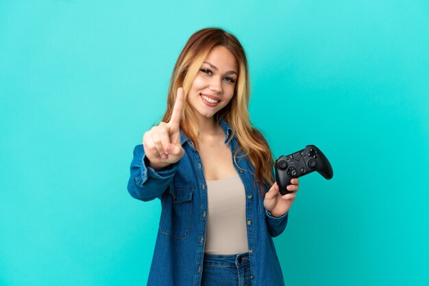 Ragazza bionda dell'adolescente che gioca con un controller per videogiochi su una parete isolata che mostra e solleva un dito