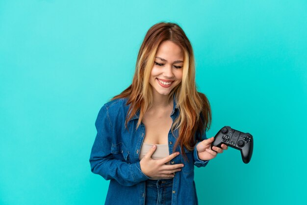 Teenager blonde girl playing with a video game controller over isolated wall smiling a lot