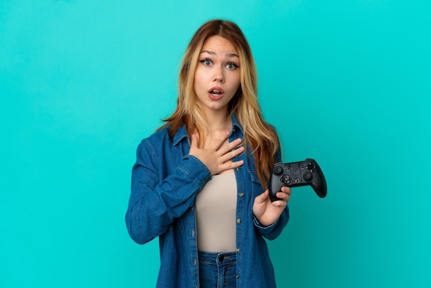 Блондинка-подросток играет с контроллером видеоигры над изолированной стеной, удивлена и шокирована, глядя вправо