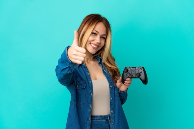 Ragazza bionda adolescente che gioca con un controller per videogiochi su un muro isolato con il pollice in alto perché è successo qualcosa di buono