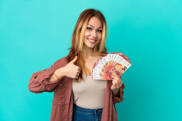 Teenager blonde girl taking a lot of Euros over isolated blue background giving a thumbs up gesture