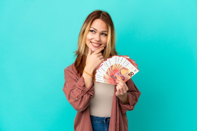 Teenager blonde girl taking a lot of Euros over isolated blue background looking to the side