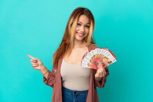 Teenager blonde girl taking a lot of Euros over isolated blue background pointing back