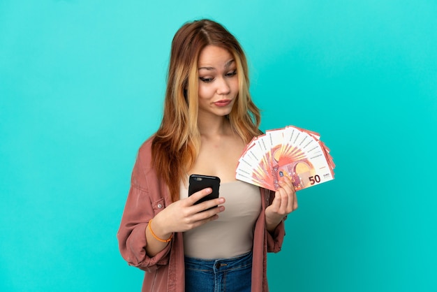 Teenager blonde girl taking a lot of Euros over isolated blue background thinking and sending a message