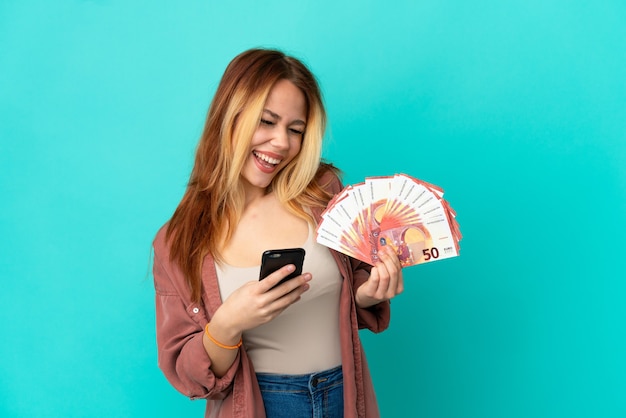 Teenager blonde girl taking a lot of Euros over isolated blue background with phone in victory position