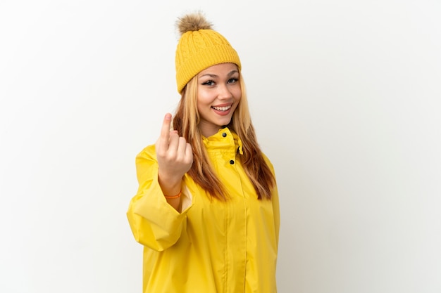 Teenager blonde girl wearing a rainproof coat over isolated white surface doing coming gesture