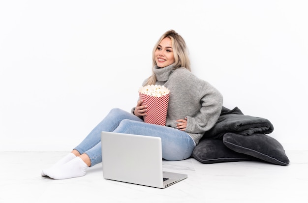 Teenager blonde woman eating popcorn while watching a movie on the laptop posing with arms at hip and smiling