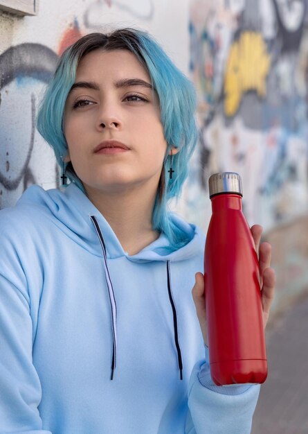 落書きの壁の近くに屋外に滞在している青い髪の十代の少女と脇に赤い水のボトルと青いパーカーのティーンエイジャー服と再利用可能なボトルのモックアップヒップスターと思春期のコンセプト