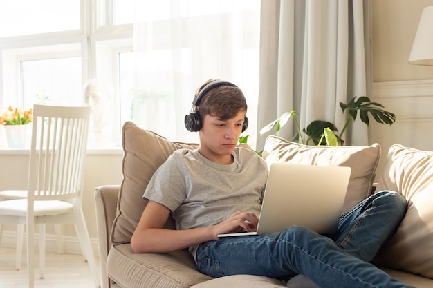 A teenager boy lying on a sofa in a room wearing black headphones on his head looks into a gray lapt