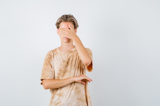 Teenager boy in t-shirt covering eyes with hand and looking scared , front view.