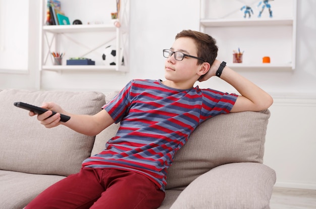 Teenager boy watching television, using TV remote while sitting on couch in living room at home