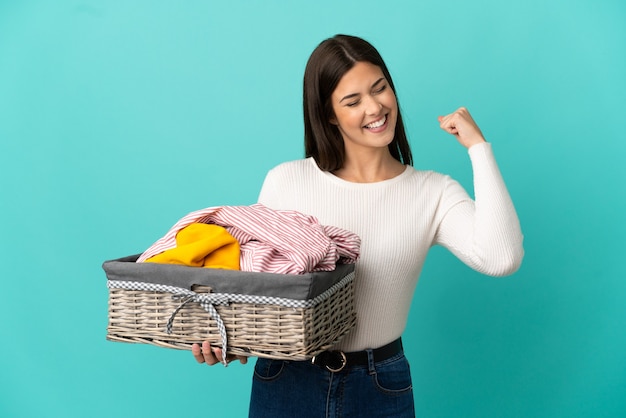 勝利を祝う青い背景で隔離の服のバスケットを保持している10代のブラジルの女の子