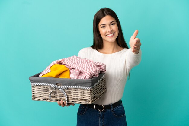 Adolescente ragazza brasiliana che tiene un cesto di vestiti isolato su sfondo blu che stringe la mano per chiudere un buon affare