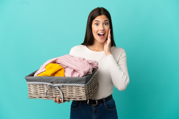 Teenager brazilian girl holding a clothes basket isolated on blue background with surprise and shocked facial expression