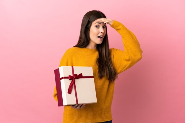 Teenager brazilian girl holding a gift over isolated pink background doing surprise gesture while looking to the side