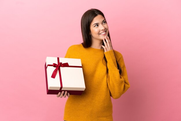 Teenager Brazilian girl holding a gift over isolated pink background looking up while smiling