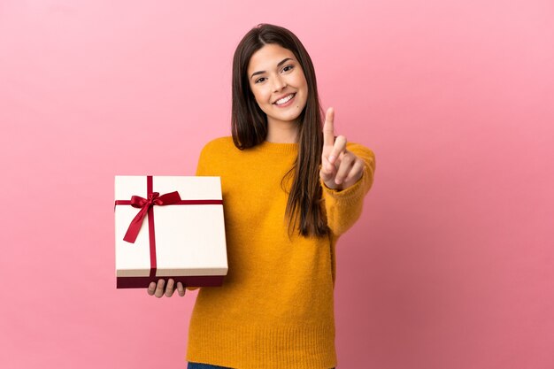 Teenager Brazilian girl holding a gift over isolated pink wall showing and lifting a finger