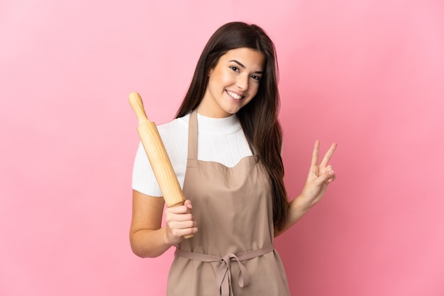 Teenager Brazilian girl holding a rolling pin isolated on pink smiling and showing victory sign