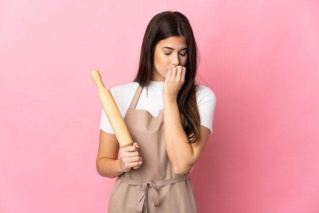 疑いを持ってピンクの壁に分離された麺棒を保持している10代のブラジルの女の子