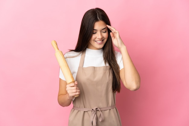 Teenager Brazilian girl holding a rolling pin isolated on pink wall laughing