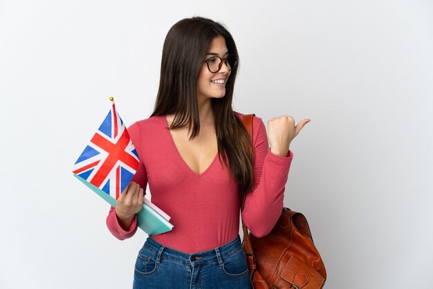 Teenager Brazilian girl holding an United Kingdom flag isolated on white wall pointing to the side to present a product